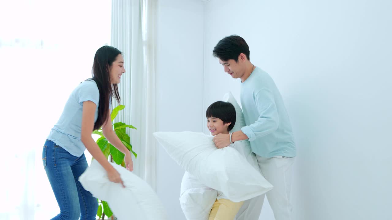 幸福的家庭时光，年轻的亚洲妈妈和爸爸在卧室的床上和儿子一起玩打枕头，笑着笑着。周末亲子时间陪孩子，好时间在家放松。视频素材