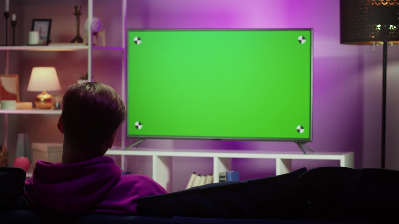 男人在客厅看电视。年轻人坐在沙发上，对着绿色的电视屏幕，背对着电视。男性使用带有色度键的智能手机。视频下载