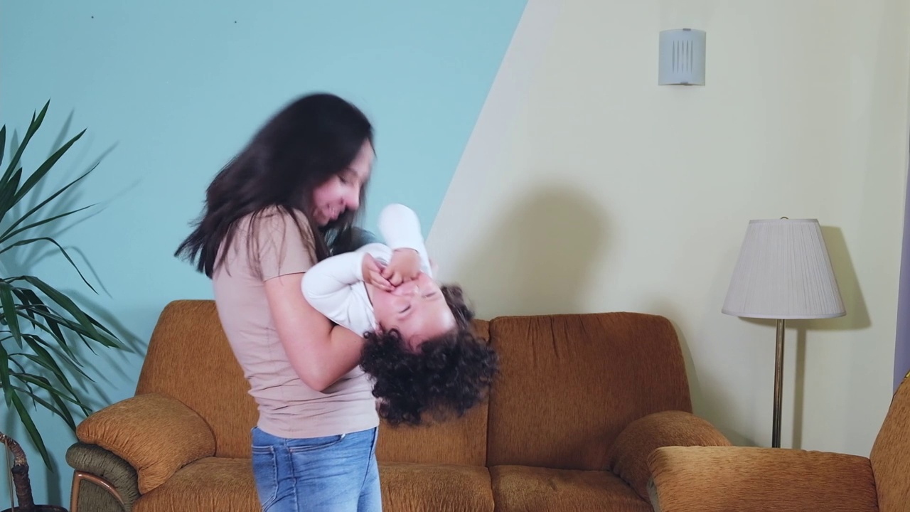 快乐的年轻成年妈妈抱着可爱的混血幼童在客厅里转来转去。视频下载