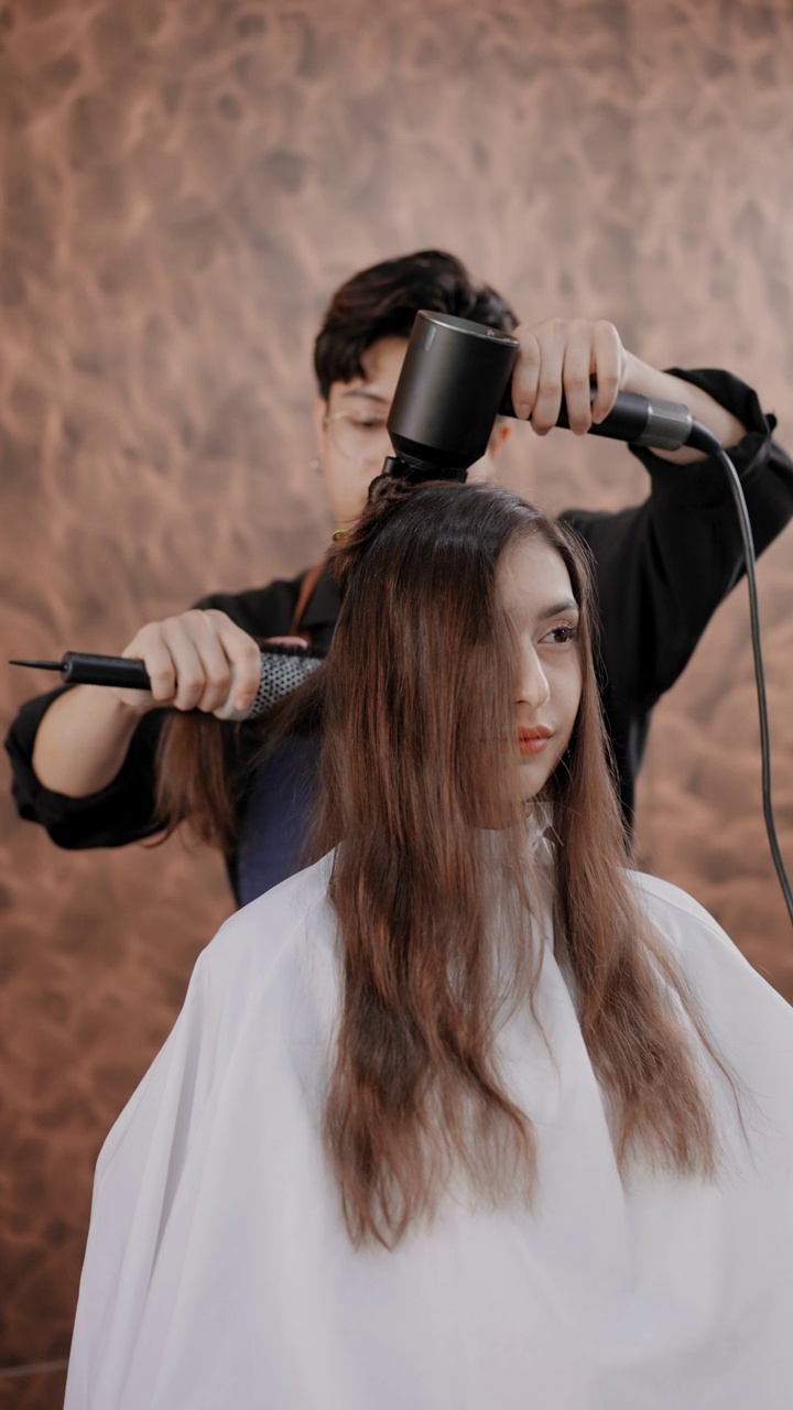亚洲男性美发师在美容院用吹风机和梳子吹干可爱女人的棕色长发。视频素材