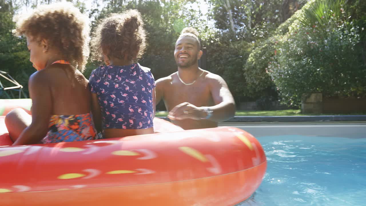 阳光明媚的夏日游泳池里，父亲在游泳池筏上为女儿们纺纱视频素材