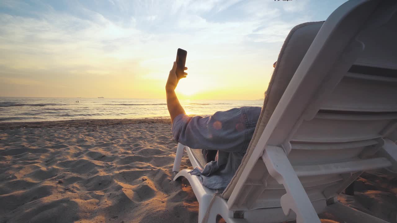 年轻英俊的男子坐在一张长椅子旁的热带沙滩上自拍，度假放松，享受日落的景色视频素材