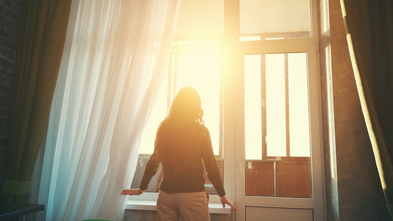 年轻女孩或长头发的妇女的后视图接近大窗户，并在夕阳的光线下打开窗帘视频素材