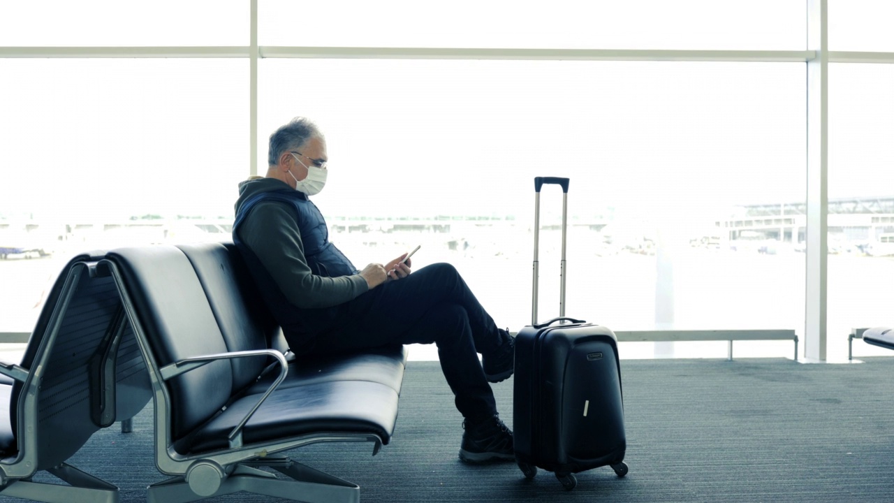 戴着防护口罩的中年男子拿着行李箱坐在机场候机厅，手里拿着手机。视频素材