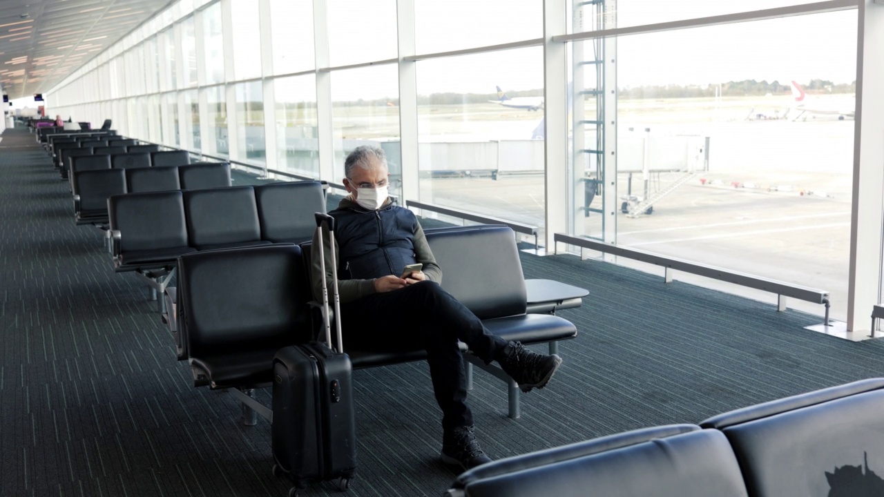 戴着防护口罩的中年男子拿着行李箱坐在机场候机厅，手里拿着手机。视频素材