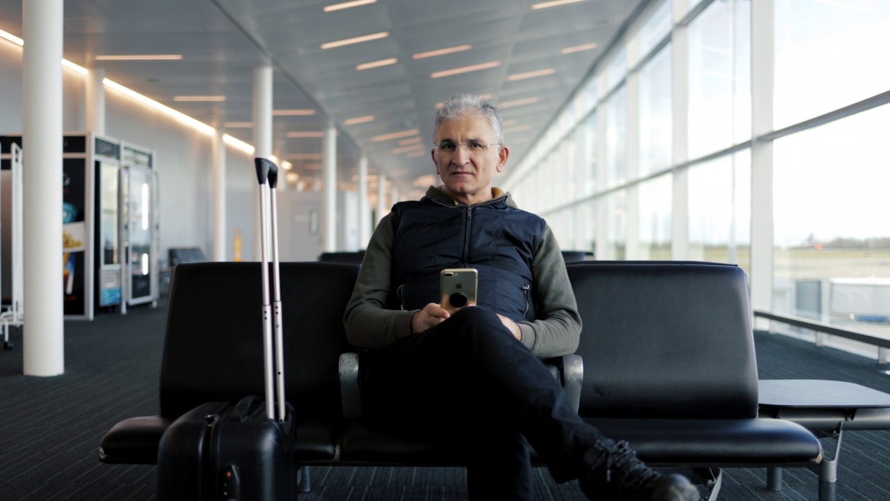 中年男子坐在机场候机大厅拿着行李，手里拿着手机视频素材