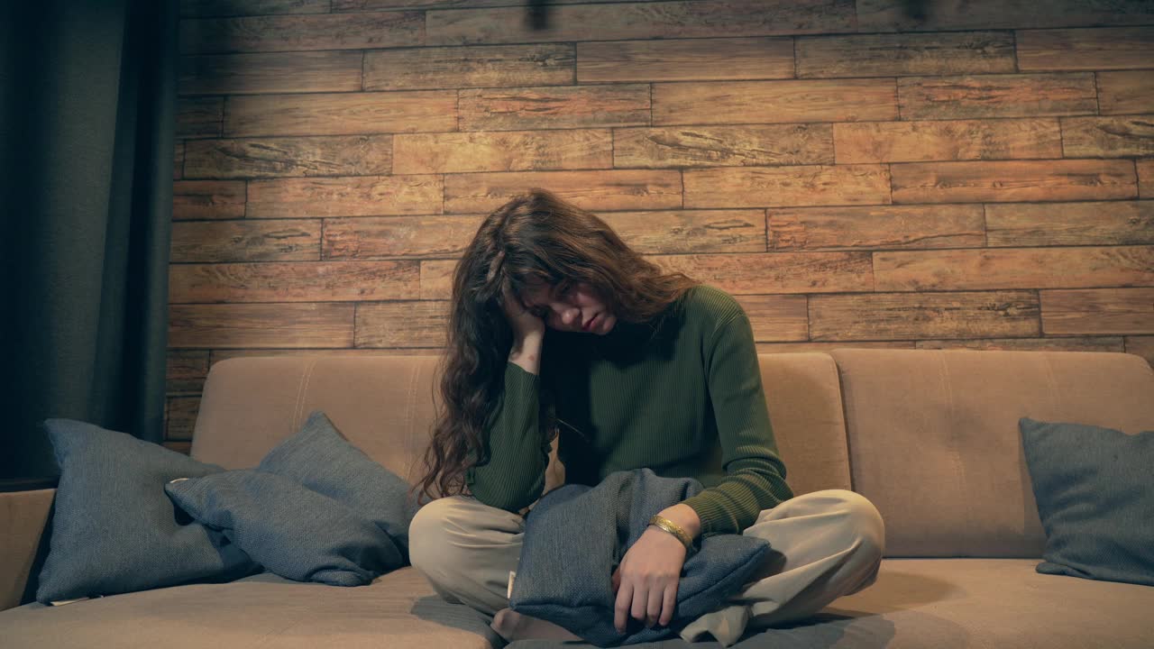 年轻悲伤的女人坐在沙发上，抚摸着自己的头和头发。抑郁症，关系问题或心理和精神问题的孤独的年轻女孩视频素材