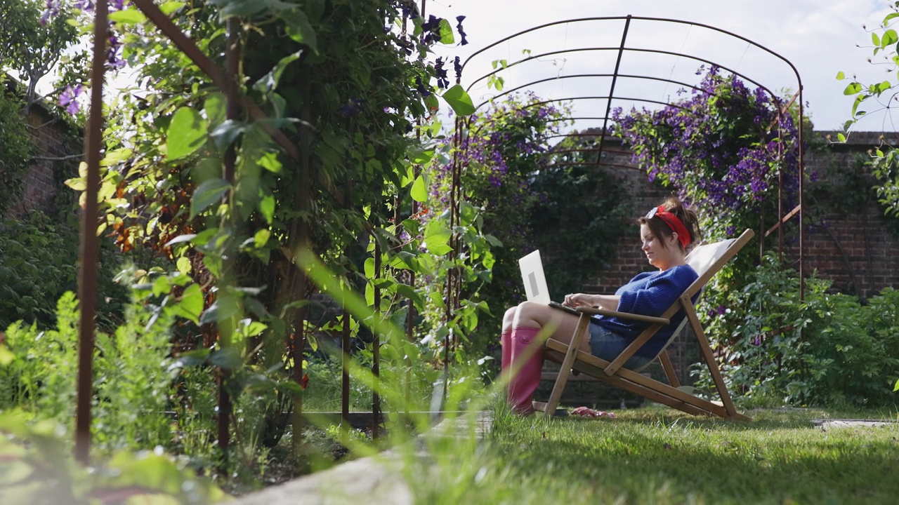 在夏日阳光明媚的后院花园里，一个女人坐在躺椅上使用笔记本电脑视频下载