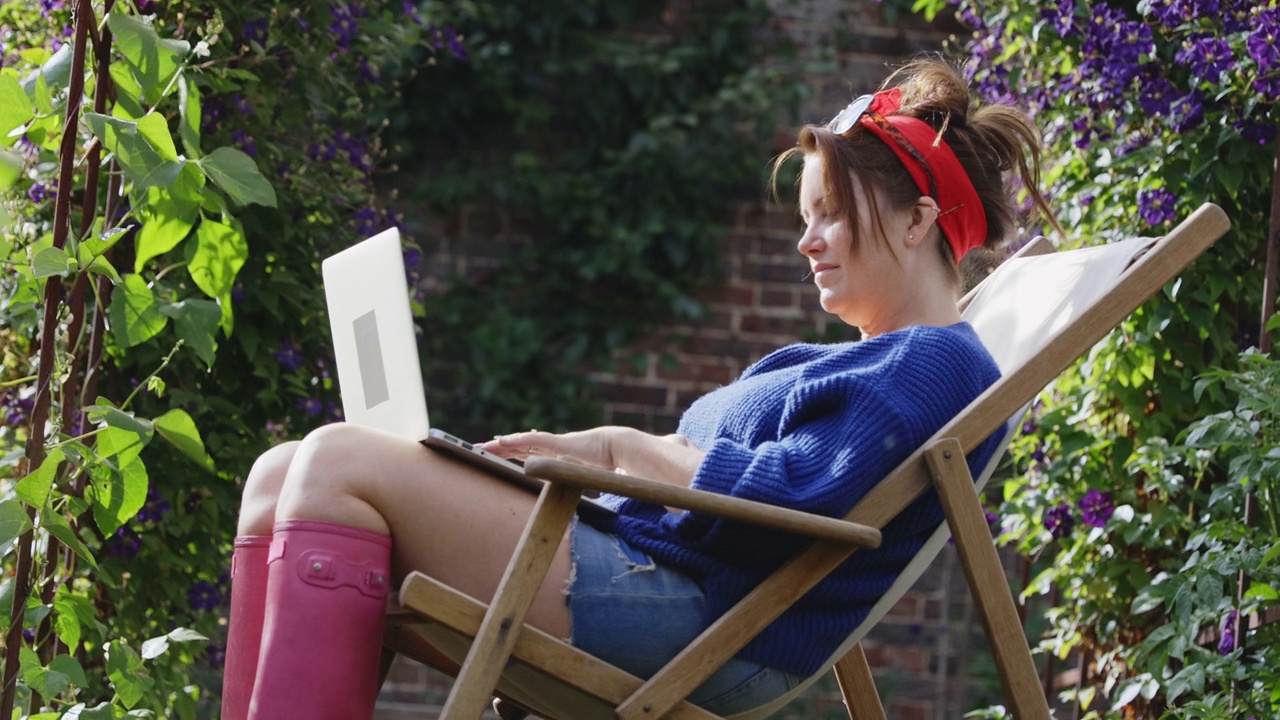一个女人在阳光明媚的夏日花园里，坐在躺椅上使用笔记本电脑视频素材