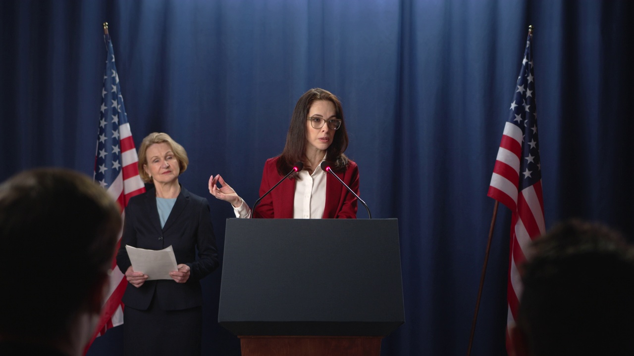 演讲期间，一名身穿红色夹克的女政治家站在蓝色幕布和美国国旗的基座后面视频素材