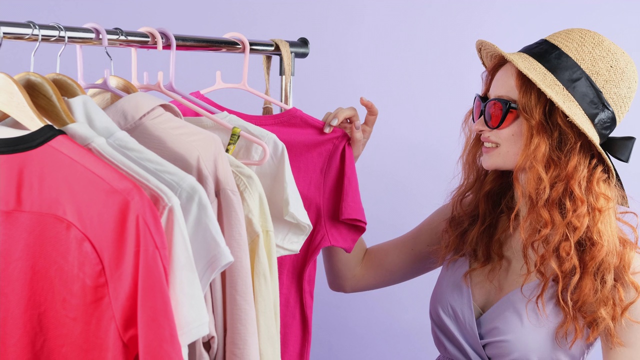 时尚的红发女人戴着太阳镜，戴着帽子，选择夏天穿的衣服，购物日的概念。视频下载