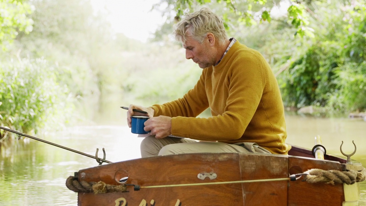成熟的男人喝着咖啡，在河上的木船里写作和钓鱼视频下载