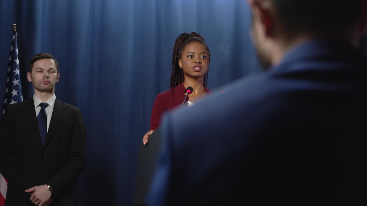 一名年轻的非洲裔美国女政治家身穿红色夹克，在印有美国国旗的蓝色幕布前发表演讲视频下载