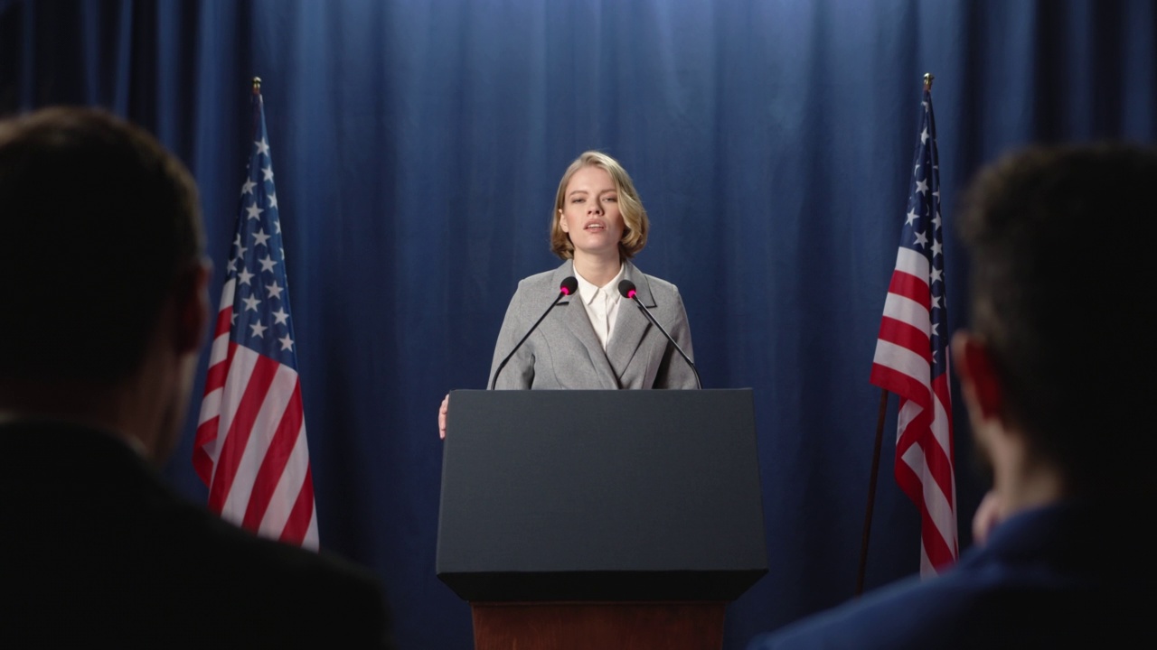 年轻的金发女政治家站在美国国旗的蓝色背景下，在辩论会上发表演讲视频下载