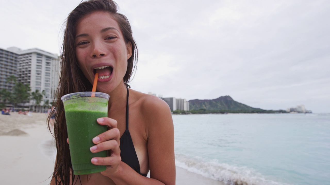 穿着比基尼在海滩上喝绿色排毒蔬菜奶昔的女人。运动女孩游泳后清新快乐。健身健康的生活方式理念与混血儿亚裔白种人女性模特。96 FPS。视频素材