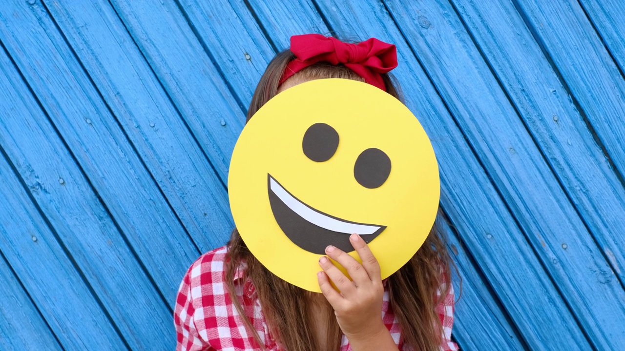 女孩躲在纸板脸后面，上面是一个快乐的笑脸表情符号。世界Emoji日。做自己的快乐视频素材