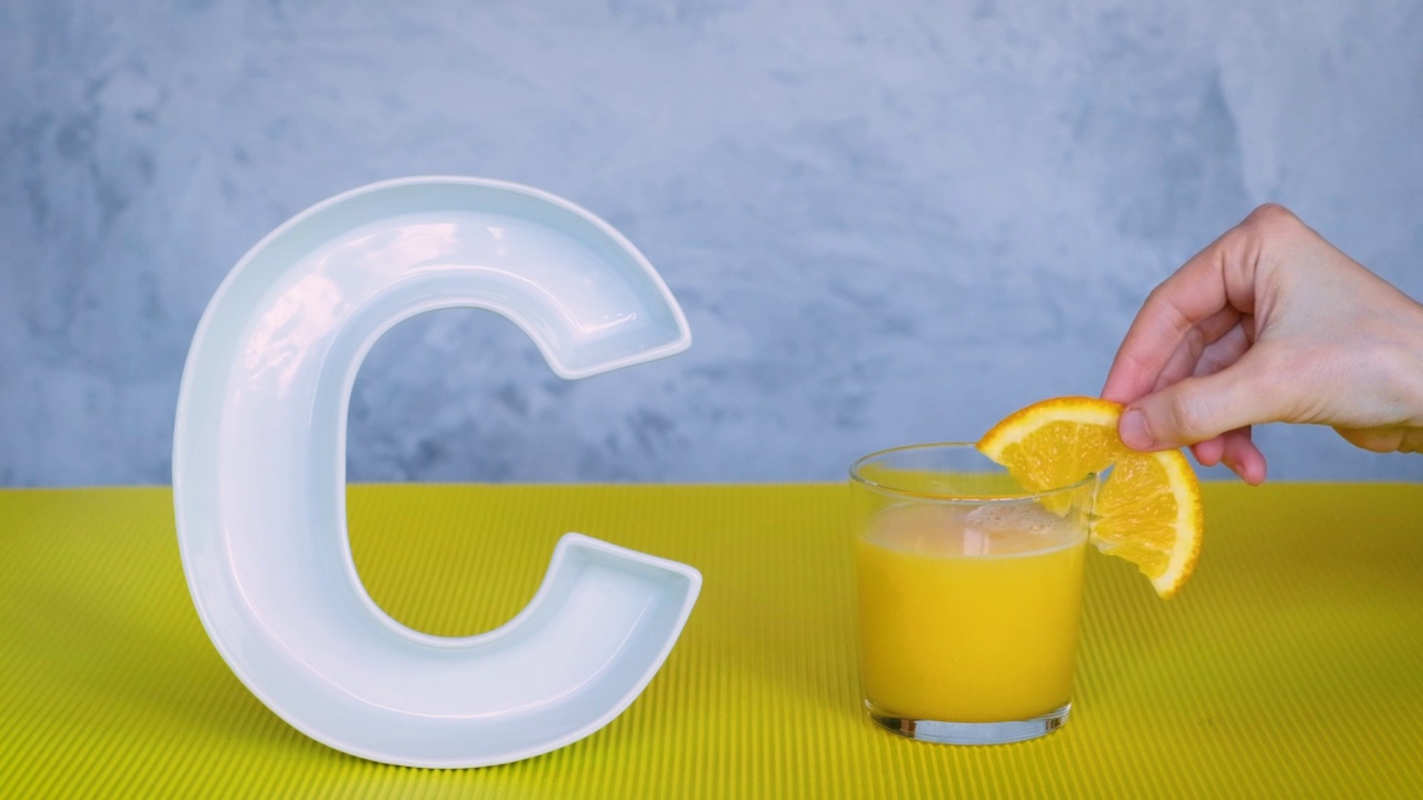 食物中的抗坏血酸或维生素C。人用手把盛有鲜榨橙汁的玻璃杯放在灰色和黄色背景的大陶瓷字母C旁边。视频下载
