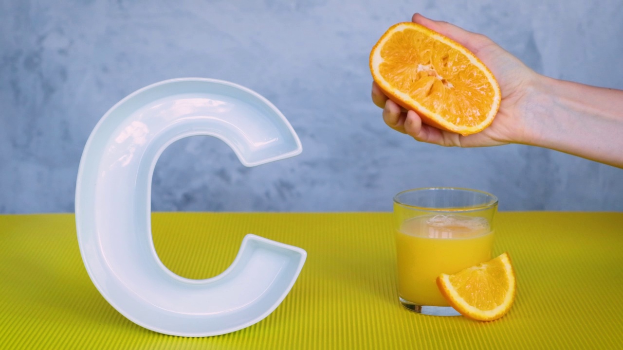 维生素C或抗坏血酸在食品中的概念。一个人用手把鲜榨的橙汁挤到一个玻璃杯里，旁边是灰色和黄色的大陶瓷字母C。视频下载