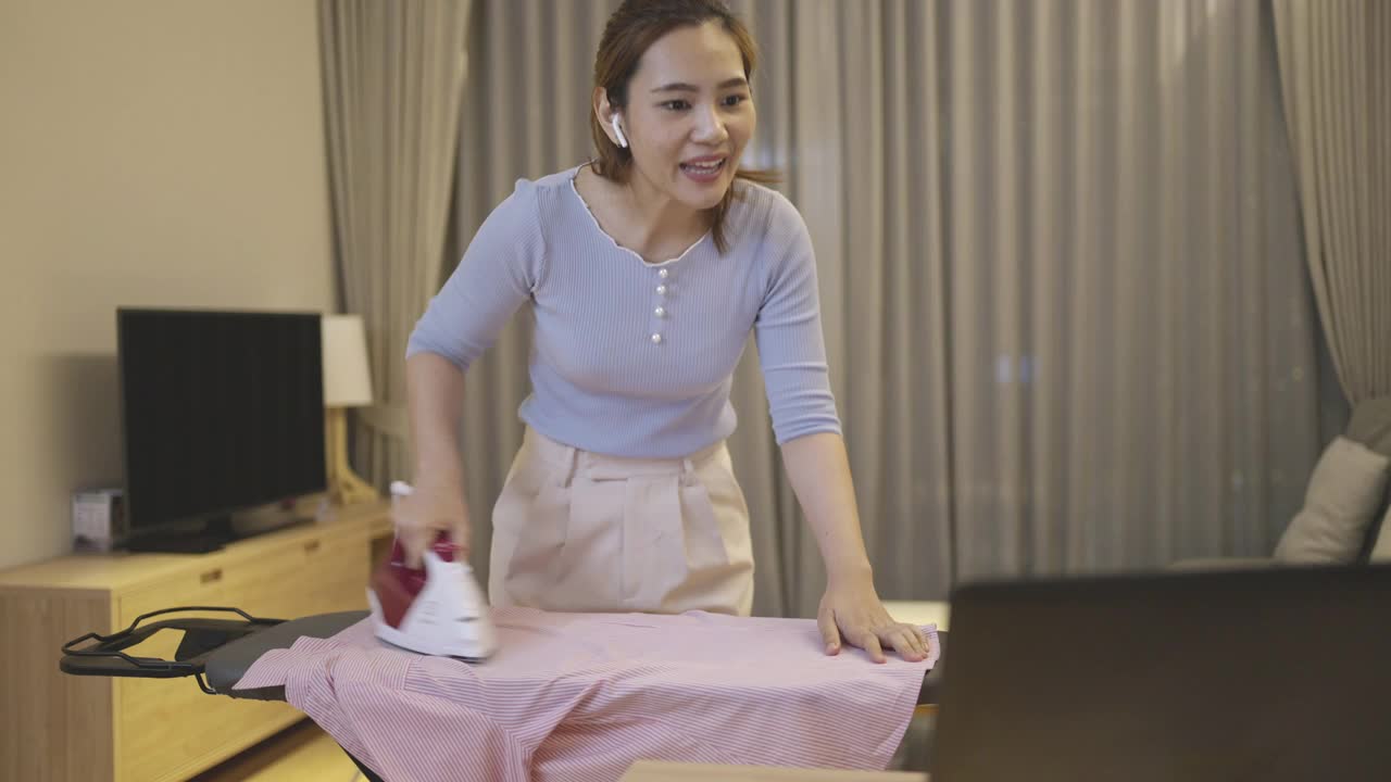 亚洲妇女在家里做家务，在客厅里熨衣服，和同事在笔记本电脑上进行在线视频会议，准备明天早上要展示的项目。视频下载