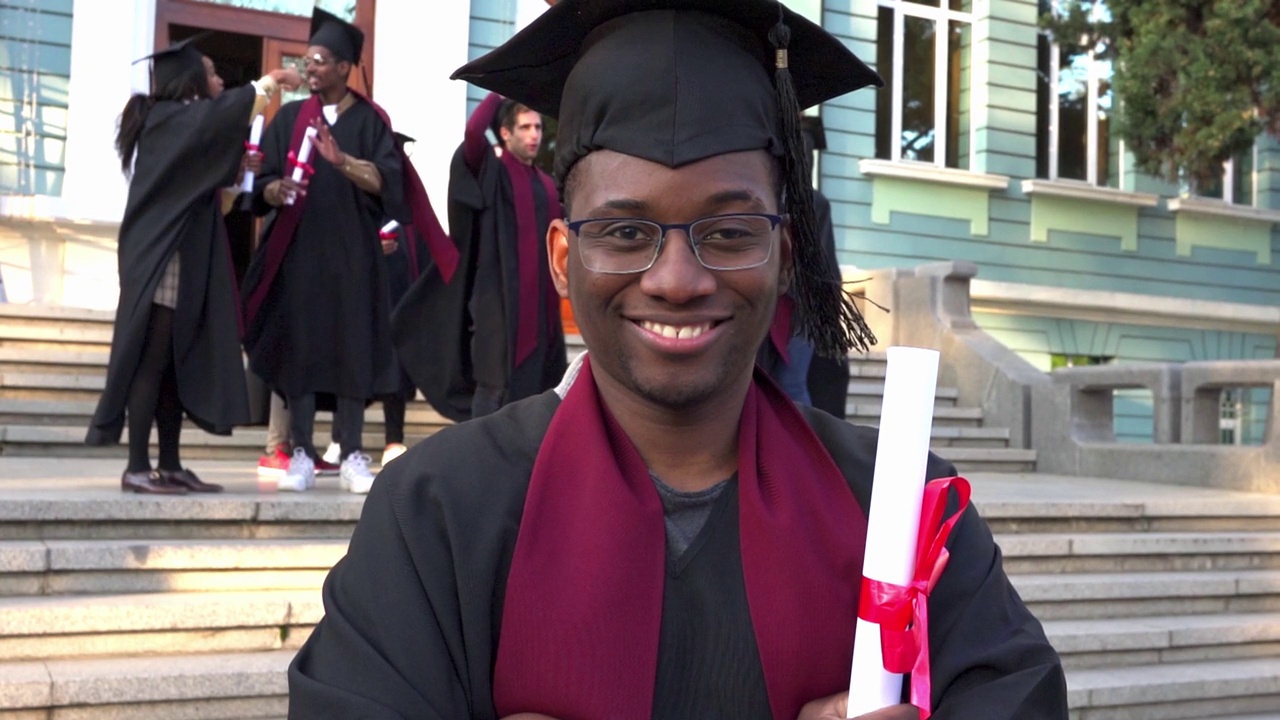 一个微笑的毕业生自豪地在大学的台阶上展示他的证书视频素材