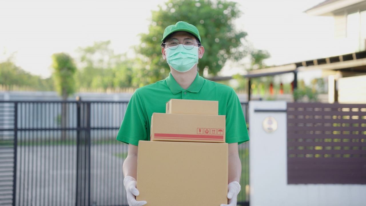冠状病毒概念，帅气的亚洲送货男子穿着绿色制服，拿着包裹纸板箱，戴着防护口罩和医用橡胶手套在客户房前。4 k决议。视频下载