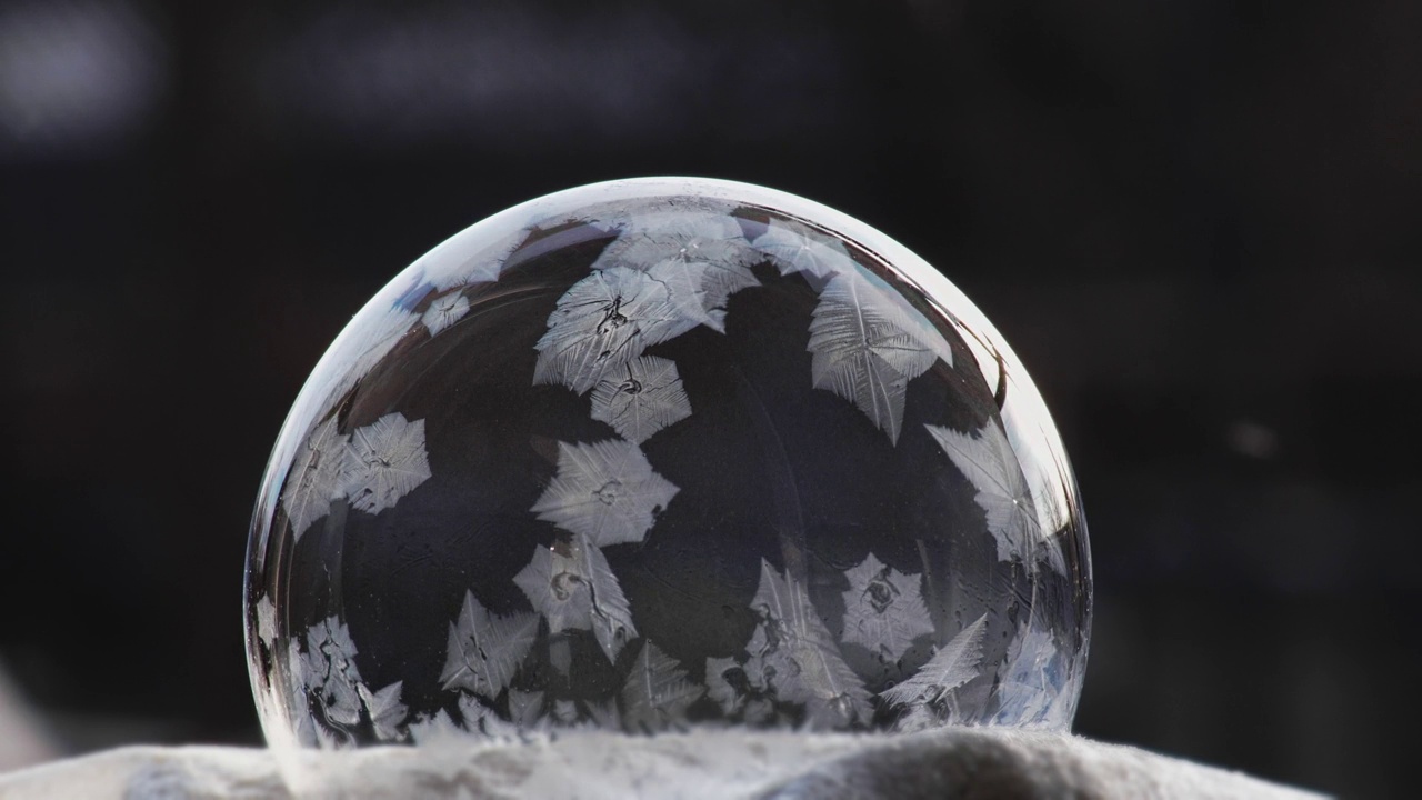 一个肥皂泡躺在黑色的雪地上，雪花落在上面结冰。冰气球模式视频下载