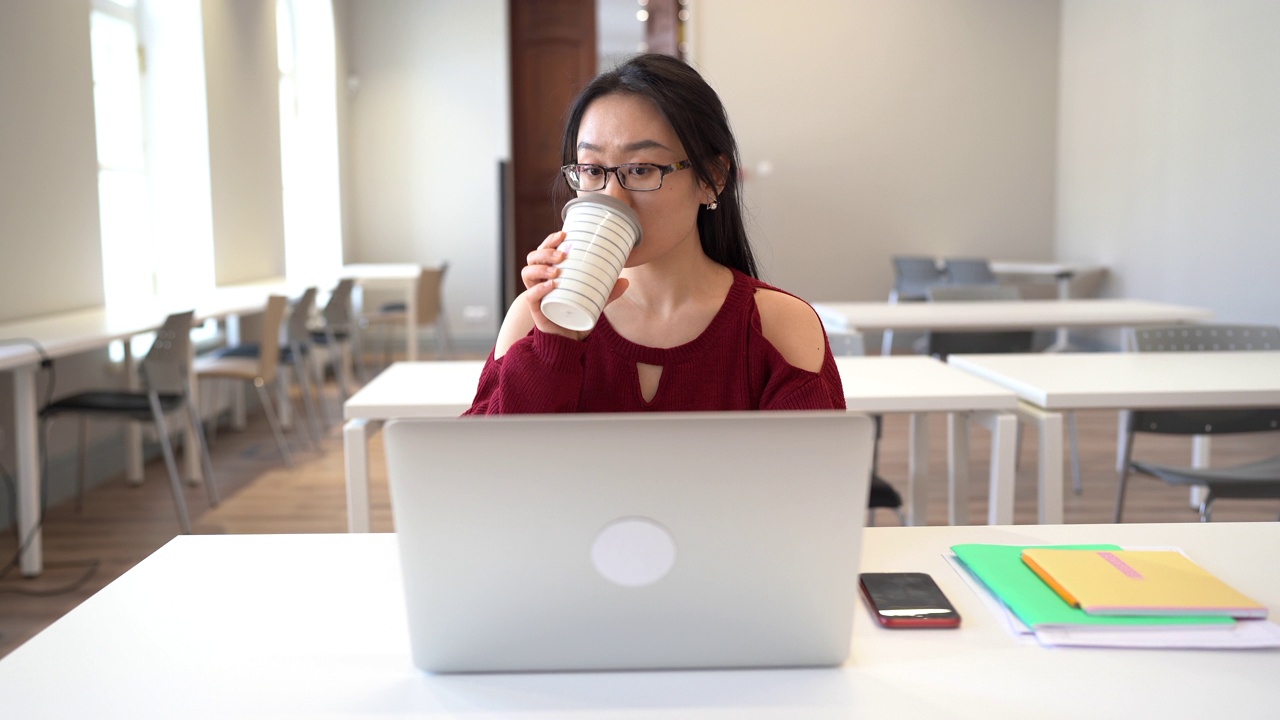 专注的亚洲女孩，戴着眼镜，坐在图书馆的桌子旁，在笔记本电脑上学习视频素材
