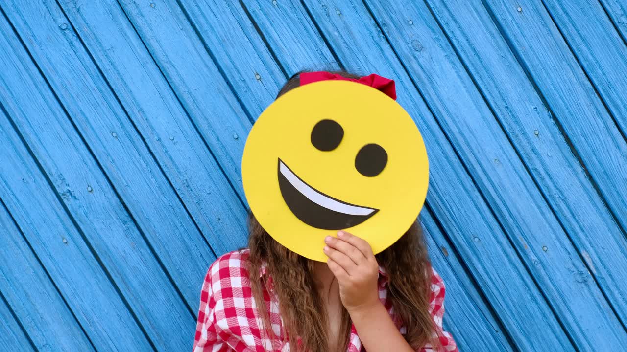 她穿着格子衬衫，头发上戴着红蝴蝶结。这个女孩把一张快乐的笑脸藏在一张纸板脸后面。世界Emoji日。做自己的快乐。用真正的微笑来代替画出来的微笑视频素材