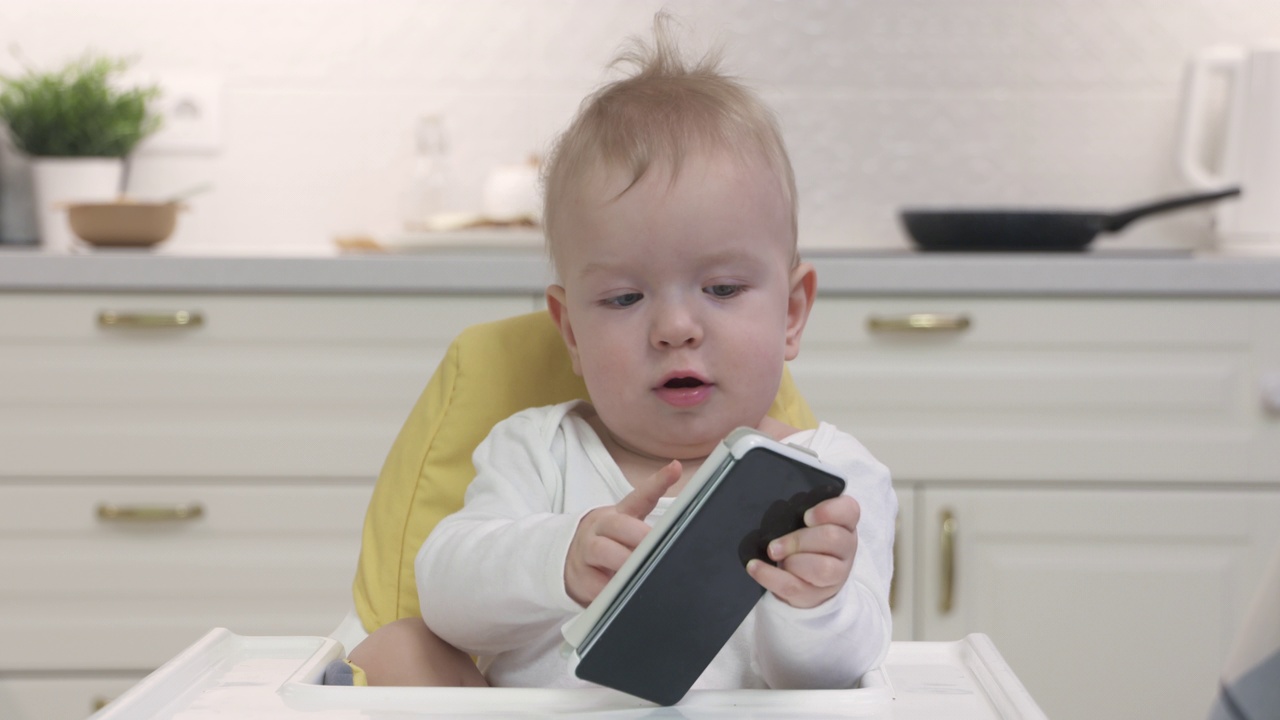 宝宝坐在高脚椅上，让宝宝玩智能手机，可爱的学步儿童拿着手机，儿童和现代科技的概念。视频素材