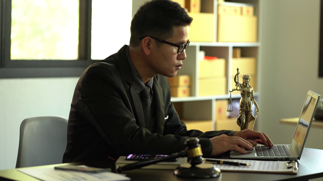一位男律师在一个木制桌子上放着黄铜天平女神的办公室里工作。司法锤与笔记本电脑法律概念和法律服务视频下载