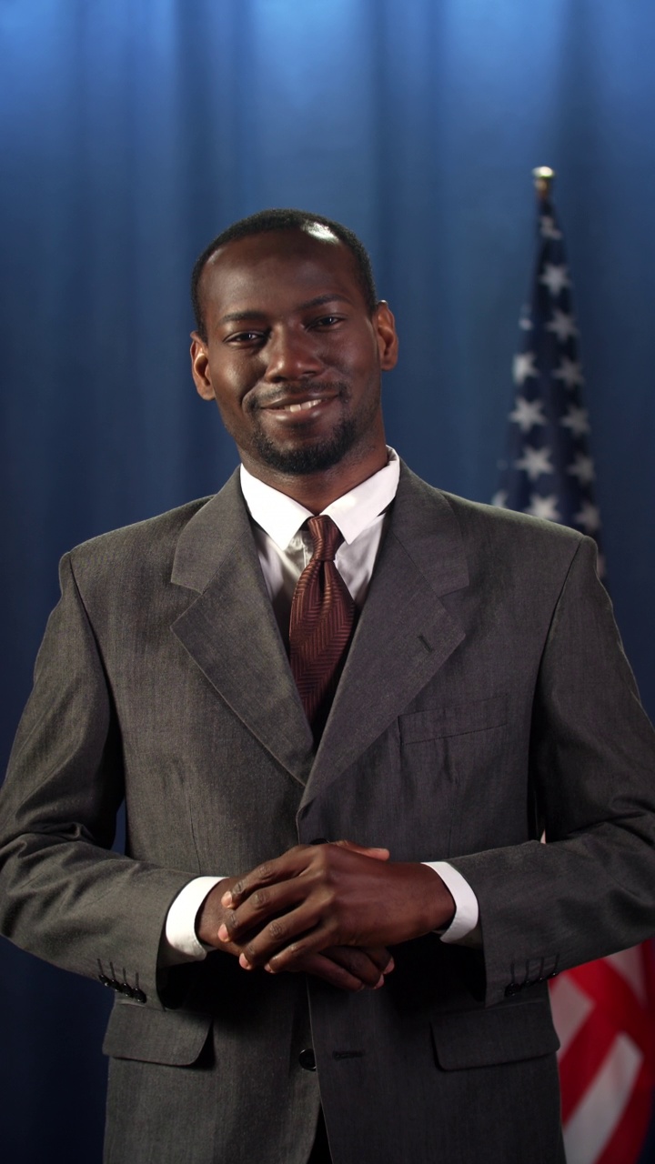 年轻的深色皮肤的政治家微笑着站在镜头前，背景是蓝色的美国国旗视频下载