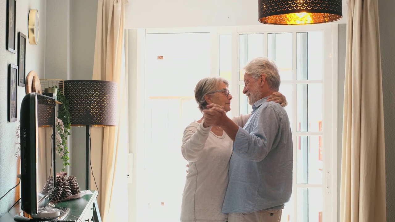 浪漫的老夫妇手牵着手在客厅里一起跳舞，幸福的老夫妇在家里跳舞庆祝。老夫妇在现代公寓里玩得很开心视频素材