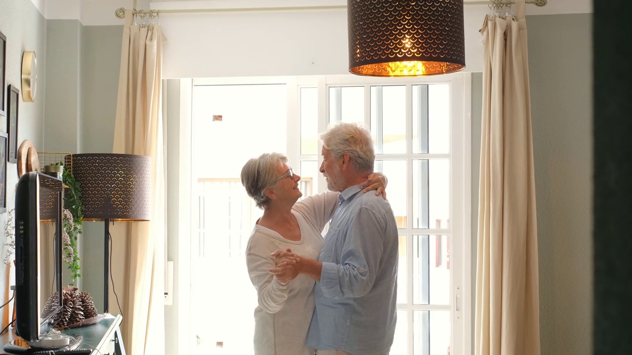 浪漫的老夫妇手牵着手在客厅里一起跳舞，幸福的老夫妇在家里跳舞庆祝。老夫妇在现代公寓里玩得很开心视频素材