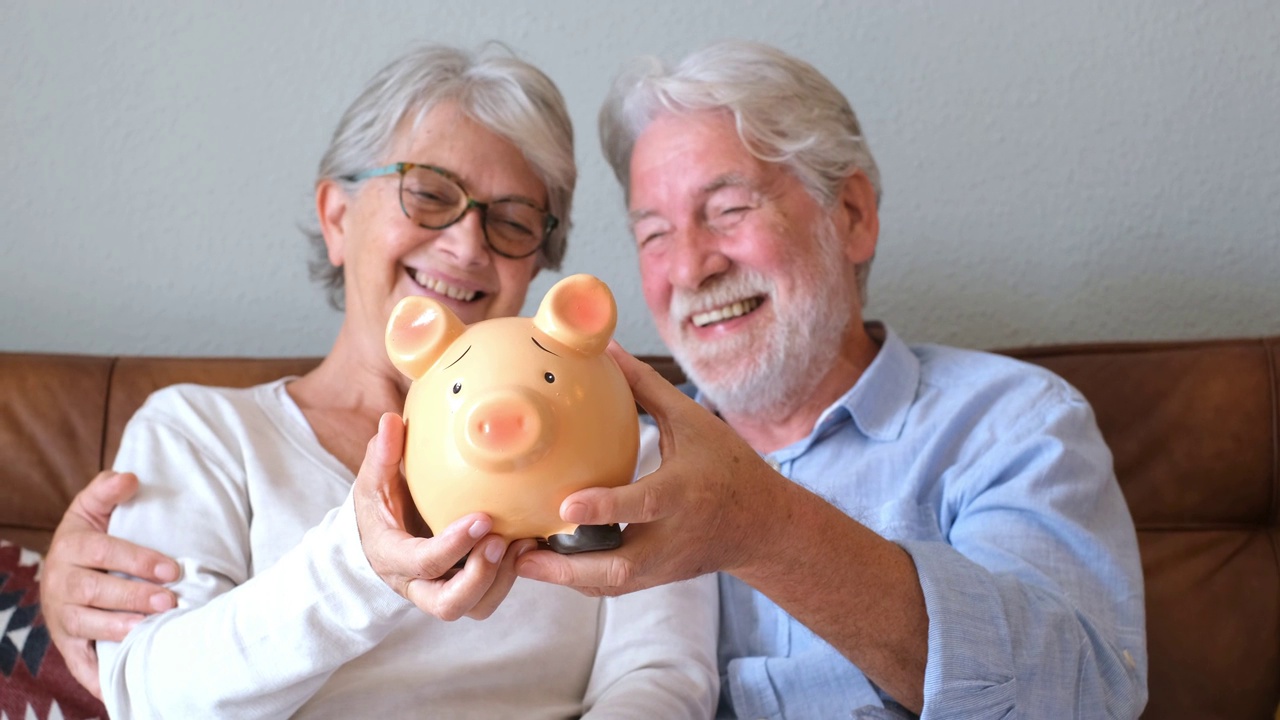 快乐的老夫妇展示储蓄罐存钱。老年男女拿着存钱罐进行投资，构思未来规划。退休的丈夫和妻子拿着硬币银行微笑视频下载