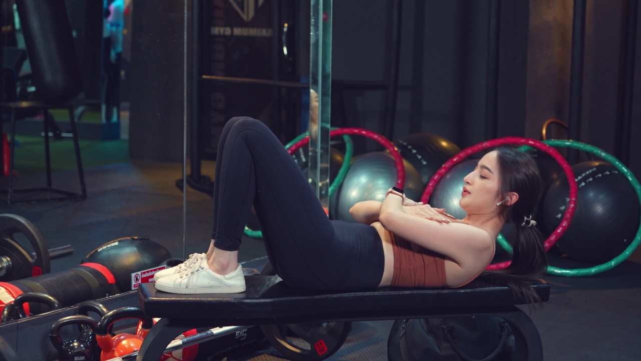 亚洲年轻女运动员在健身椅上做仰卧起坐。健身女士做腹肌仰卧起坐。在健身房做腹肌锻炼。视频素材