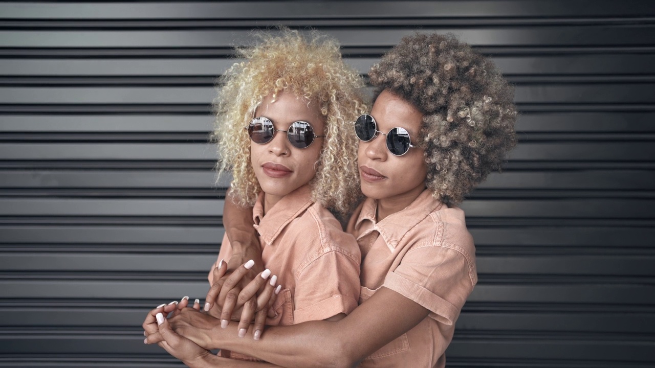 年轻的黑色双胞胎姐妹的肖像金发和太阳镜，微笑和拥抱彼此的爱和感情视频素材