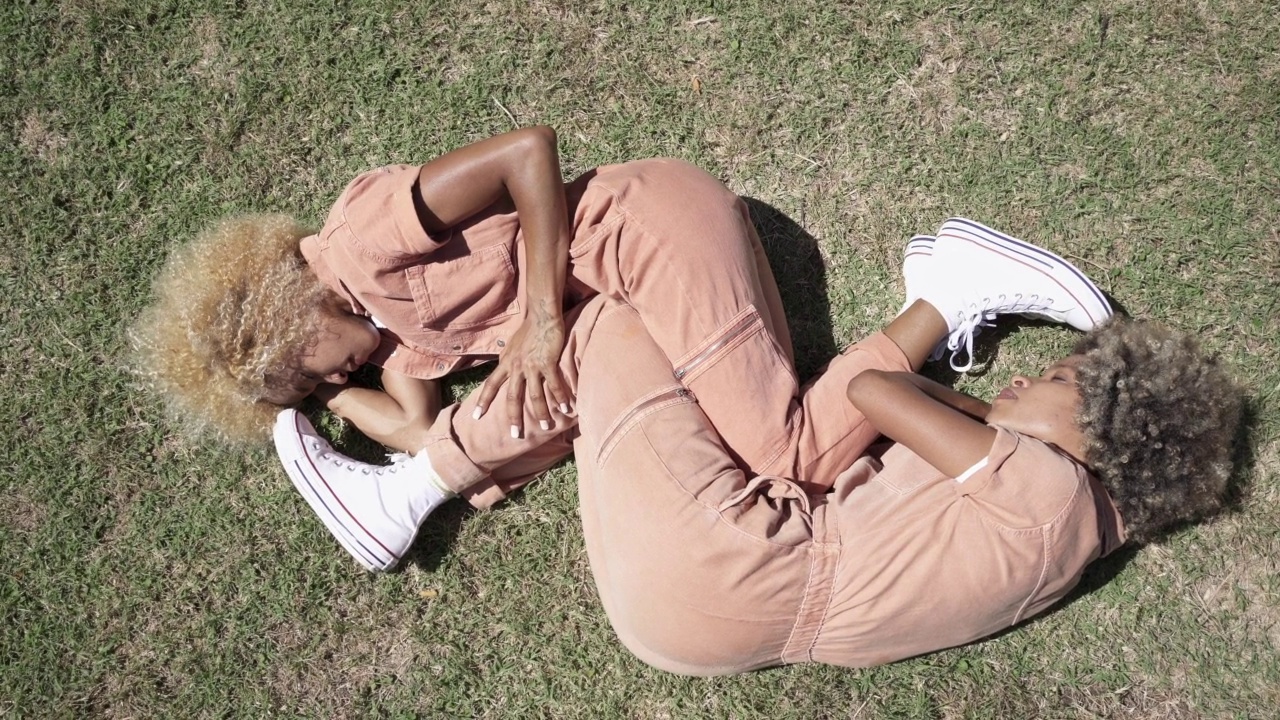 非裔美国双胞胎姐妹躺在草地上，以胎儿的姿势睡在一起。家庭中的情感联系视频素材