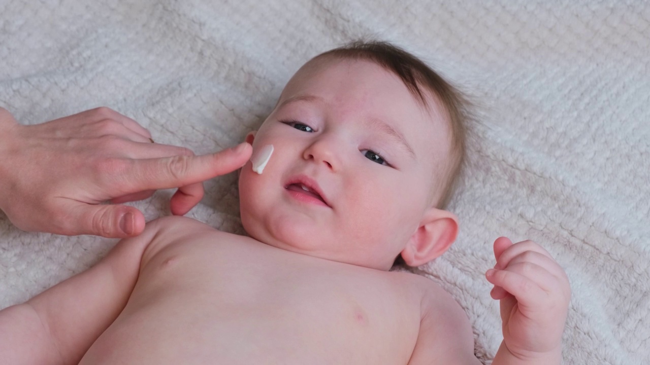 妈妈的手正在给一个快乐的婴儿涂抹乳霜。妈妈在沙发上按摩一个八个月大的微笑的蹒跚学步的孩子视频下载