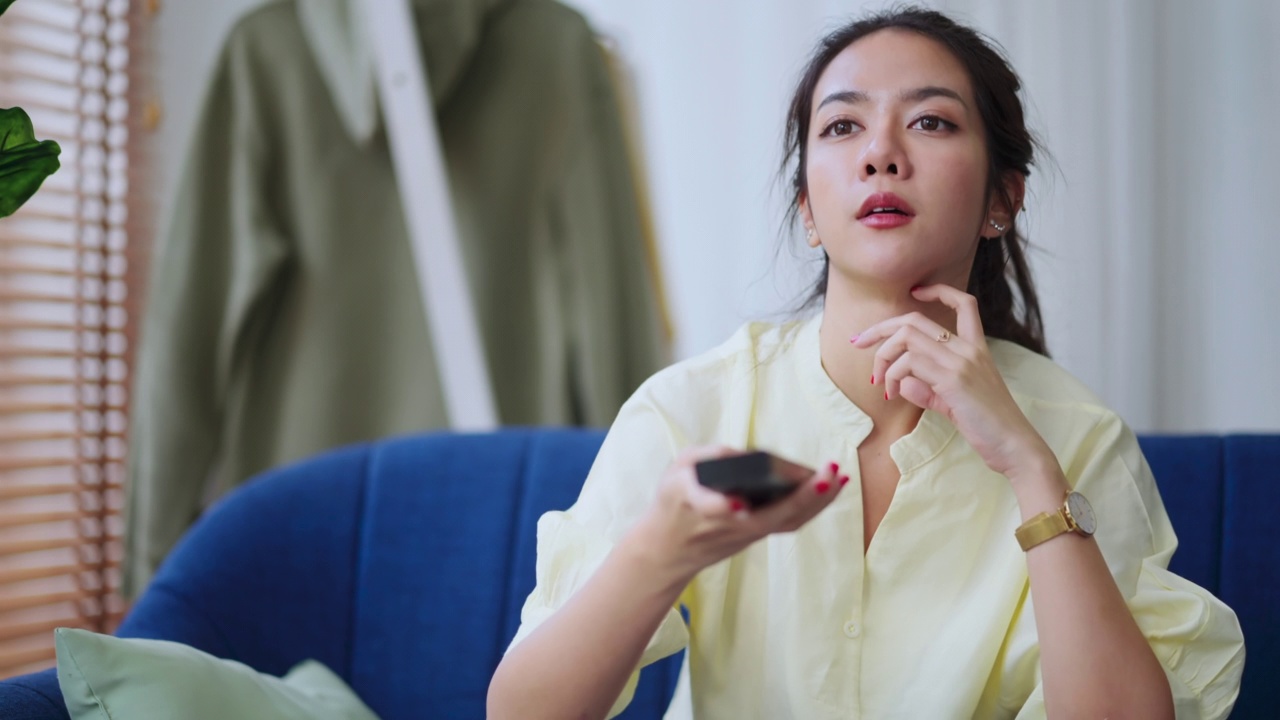 出生的亚洲女性换电视节目频道在客厅的沙发上找到她感兴趣的节目，亚洲女性用手用遥控器换电视频道带着无聊和尝试的表情视频素材