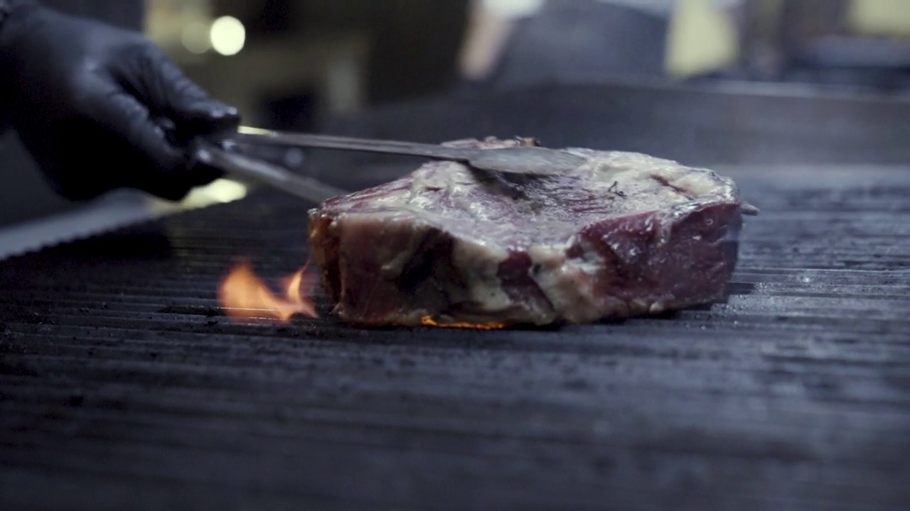 多汁的牛排在炭火上烧烤，黑色的背景，在火焰的烟。晚上烧烤时，厨师用钳子把肉放在烤架上。用炽热的火焰做晚餐。视频素材