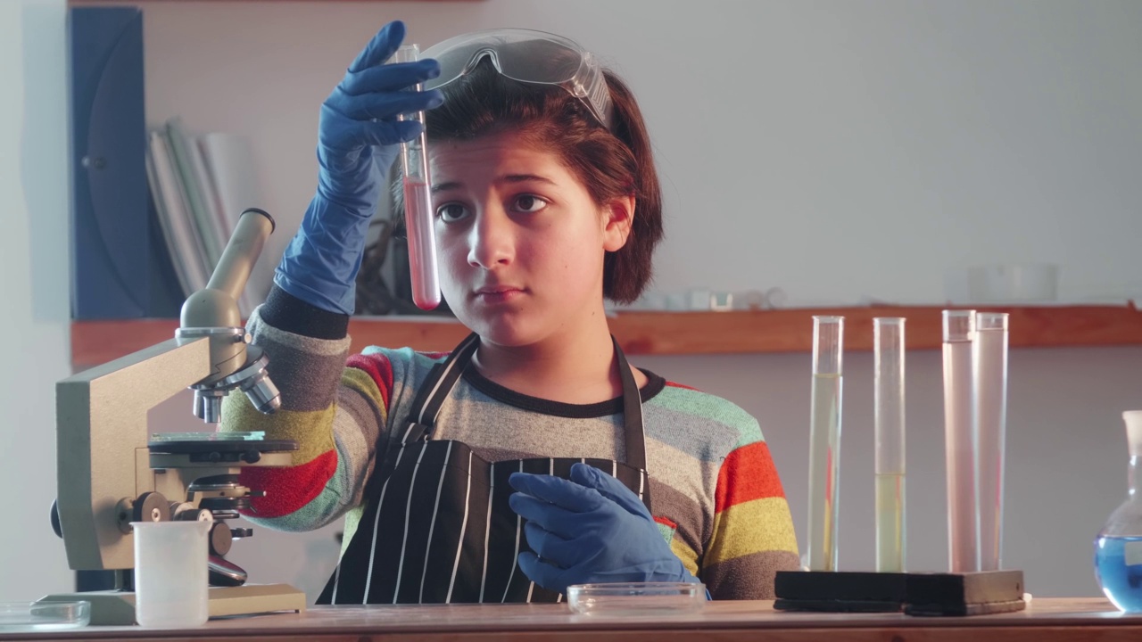 年轻的科学家。一个可爱的十几岁的男孩戴着护目镜和手套，在实验室设备和显微镜的背景下看着试管中的粉红色液体。视频素材