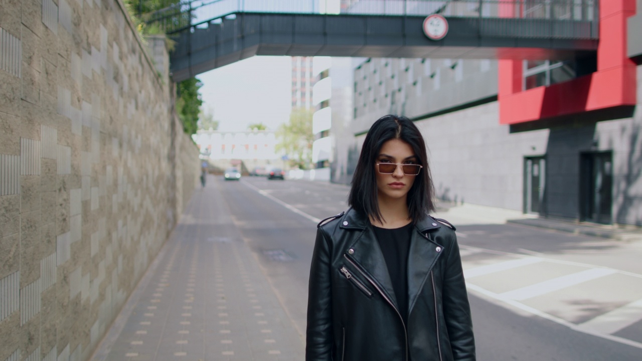 自信诱人的黑发女性穿着黑色皮夹克太阳镜站在户外城市街道看着相机在夏天的一天调情。都市风格时尚，肖像美丽性感的女人视频素材