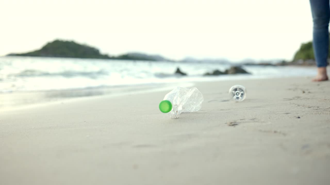 近距离拍摄，女子手捡起沙滩上的塑料瓶。女志愿者在沙滩上清理垃圾，让大海变得美丽。世界环境日的概念。拯救世界。视频素材