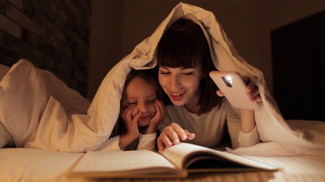 年轻漂亮的妈妈和可爱的女儿在床上的毯子下用手机手电筒看书视频素材