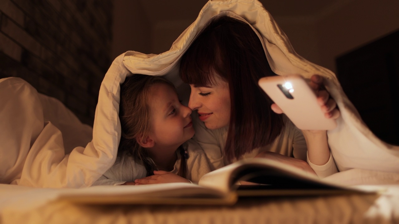 晚上，妈妈和可爱的女儿盖着毯子躺在床上读童话故事视频素材