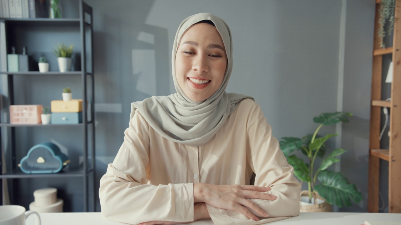 年轻貌美开朗的亚洲穆斯林女商人，戴着头巾，随意地坐在书桌上，看着摄像机，远程工作视频，笔记本电脑在线电话在家里的客厅。伊斯兰女孩的生活方式概念。视频素材