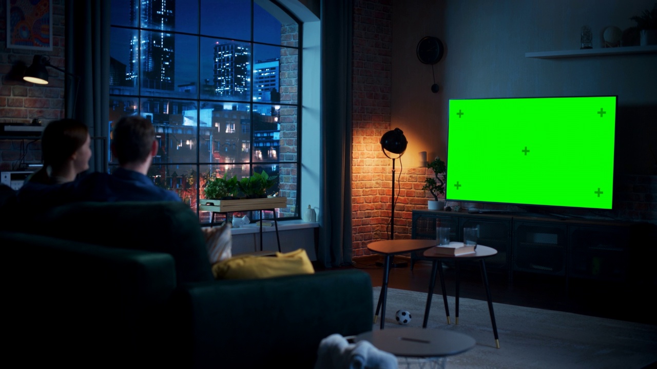 年轻夫妇花时间在家里，坐在沙发上看电视，绿色屏幕模拟显示在他们时尚的阁楼公寓。男人和女人观看晚间电影，吃爆米花。视频下载
