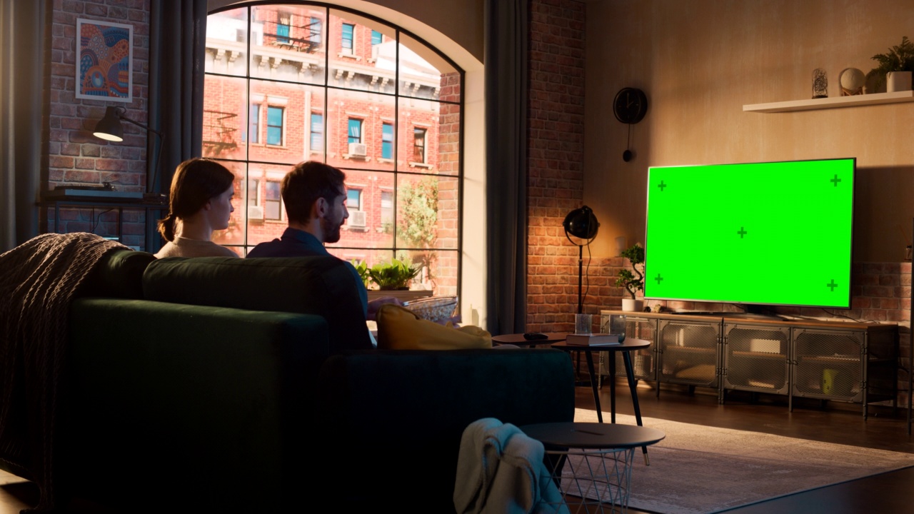 年轻夫妇花时间在家里，坐在沙发上看电视，绿色屏幕模拟显示在他们时尚的阁楼公寓。男人和女人流媒体电影或表演和吃爆米花。视频下载
