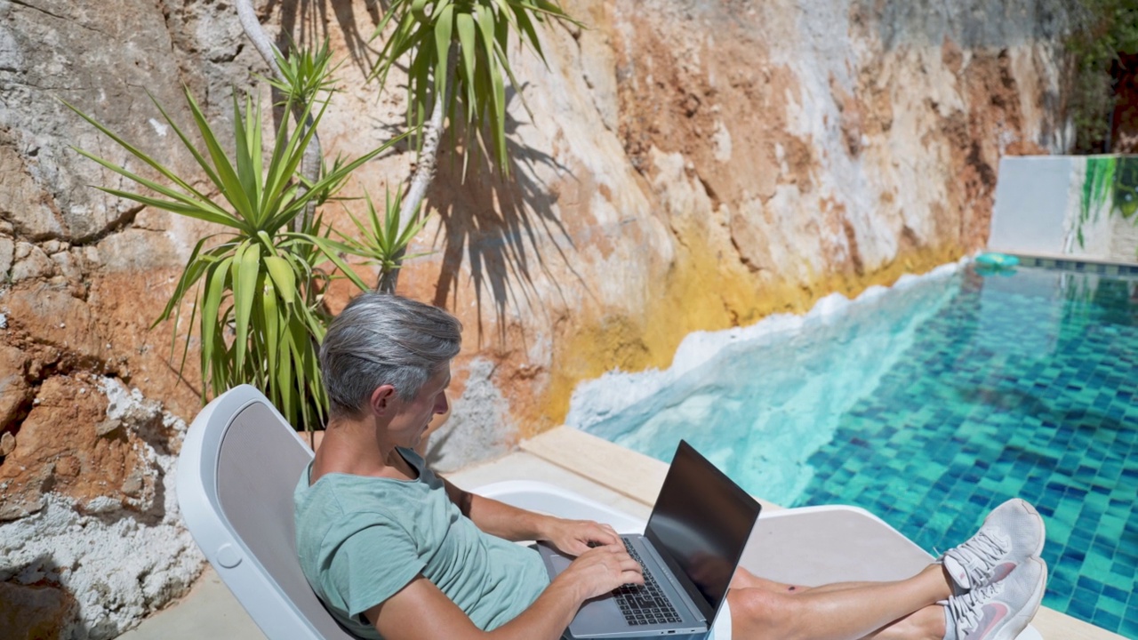 侧视图放松的成人男子在度假酒店的游泳池冷却，在笔记本电脑上看新闻或视频空屏幕视频素材