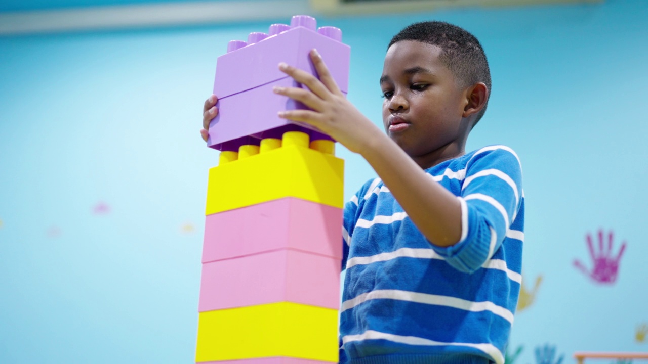 非洲黑人小孩，一个7岁的男孩试图把彩色的塑料积木尽可能高的连接起来，同时他需要注意和保护积木不被他可爱的妹妹跑和玩。视频下载